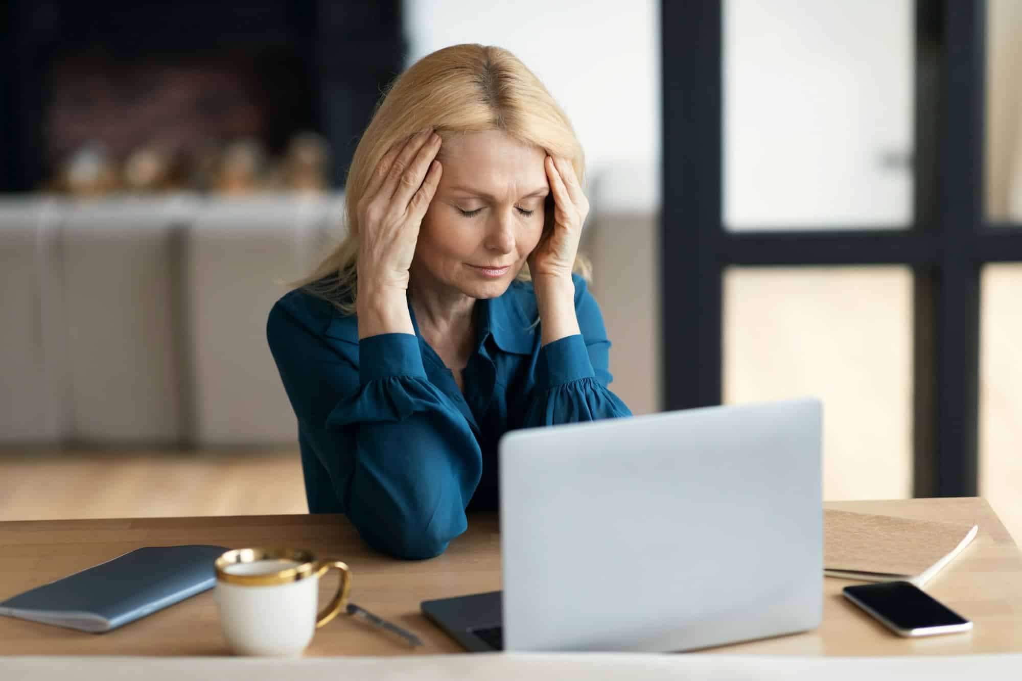 Sad mature lady sitting at table in front of computer, suffering from headache, menopause, stress in